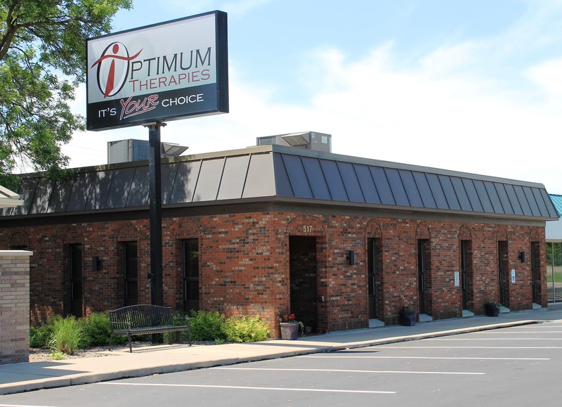 A single-story brick building with large windows and a sign reading Optimum Therapies – Its Your Choice. The building has a parking lot and a small bench near the entrance.