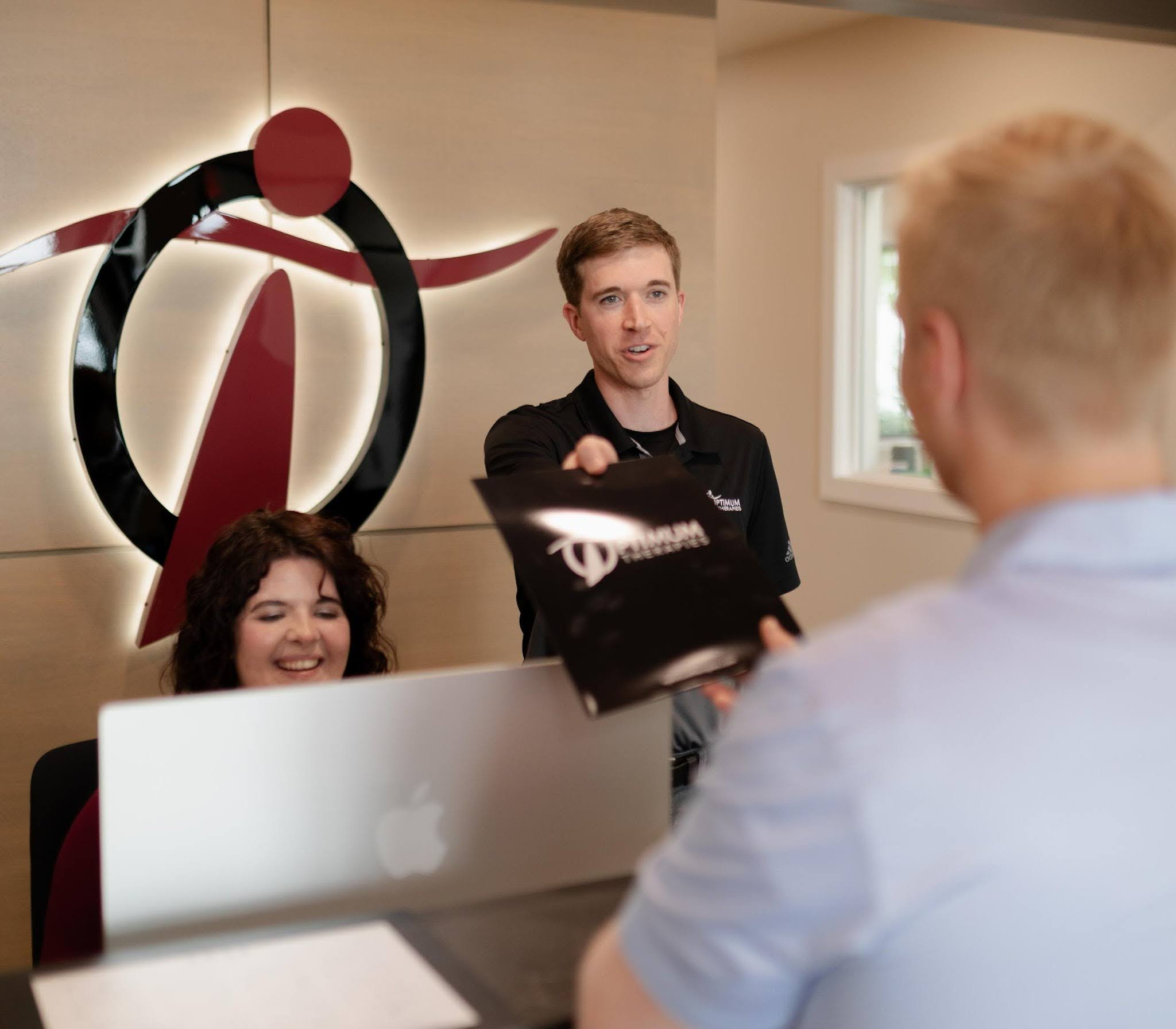 A man stands behind a reception desk handing a black folder to a blonde man, while a woman sits at the desk smiling. A stylized logo featuring a red and black figure is displayed on the wall behind them.