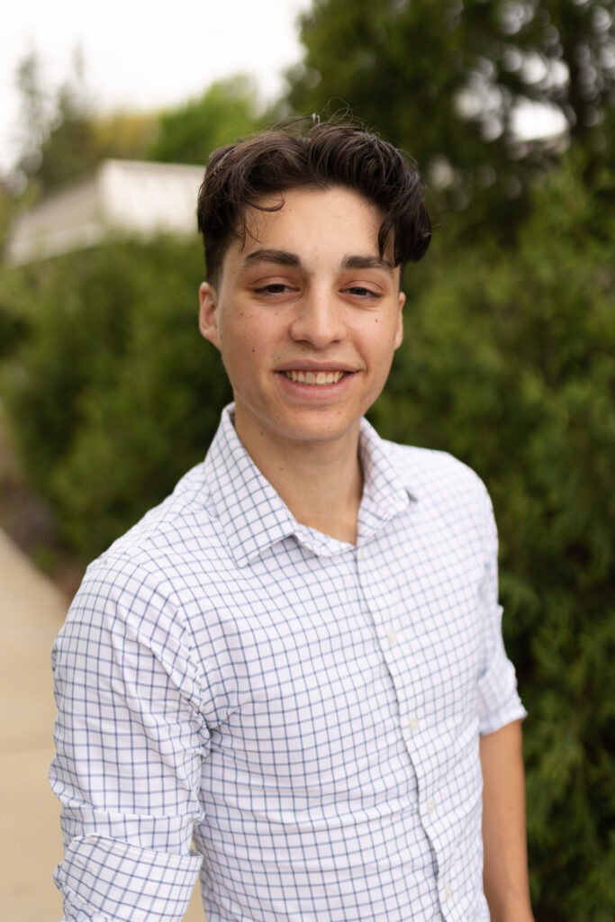 A young man with short dark hair, wearing a white dress shirt with blue checks, stands outdoors on a sidewalk with greenery and blurred trees in the background, smiling at the camera.