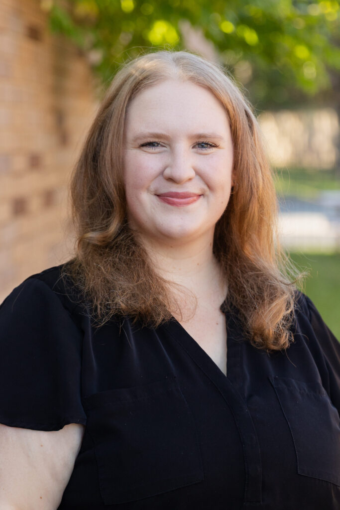 A woman with fair skin and wavy, light brown hair smiles at the camera. She is wearing a black top and standing outdoors with a blurred background of greenery and a brick wall.
