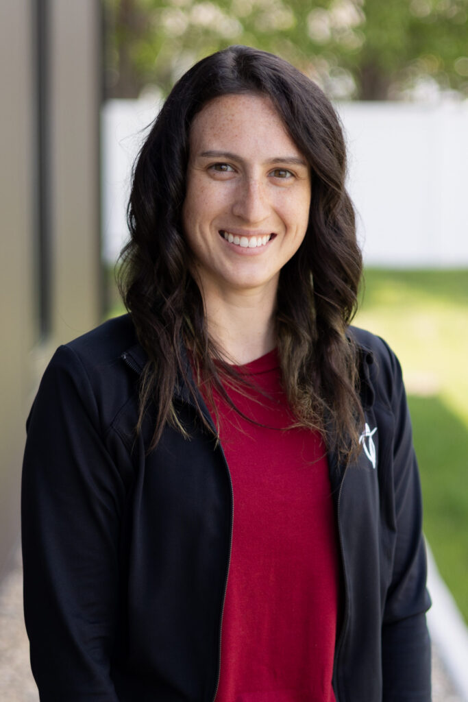A woman with wavy brown hair, wearing a red shirt and a black jacket, smiles while standing outdoors. There is greenery and a white fence blurred in the background.