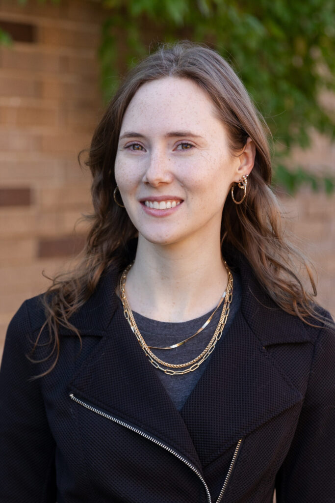 A woman with wavy brown hair wearing gold hoop earrings, layered gold necklaces, and a black jacket stands outside in front of a brick wall and greenery, smiling softly at the camera.