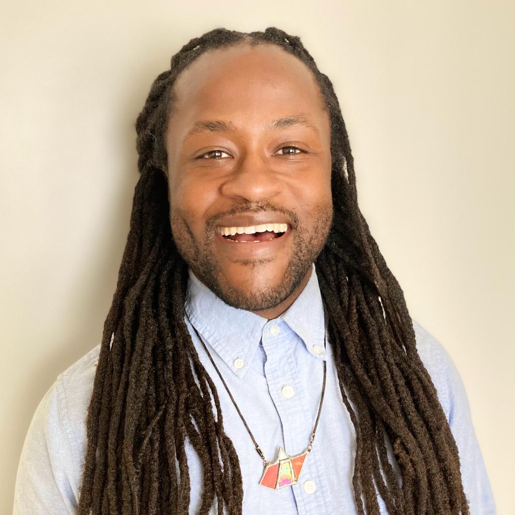 A smiling person with long, dark dreadlocks wears a light blue button-up shirt and a necklace with a colorful pendant, standing against a plain light background.