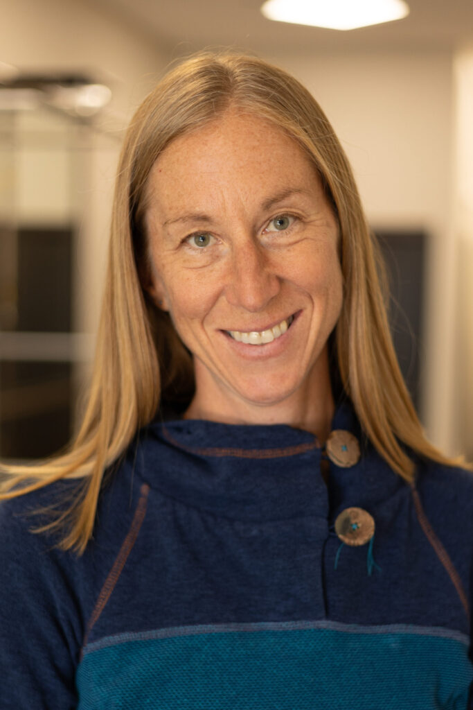 A woman with long blonde hair and wearing a blue sweater with large buttons smiles warmly at the camera indoors, with a softly blurred background.