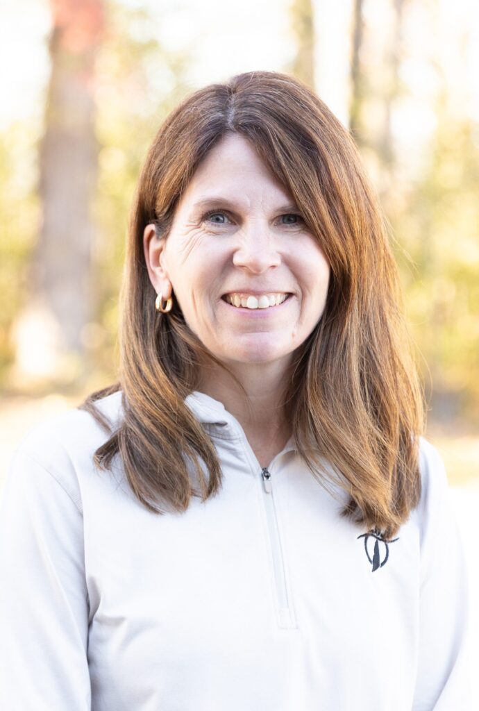 A woman with straight, shoulder-length brown hair smiles outdoors. She is wearing a light-colored quarter-zip top and gold hoop earrings, with a background of blurred trees and sunlight.