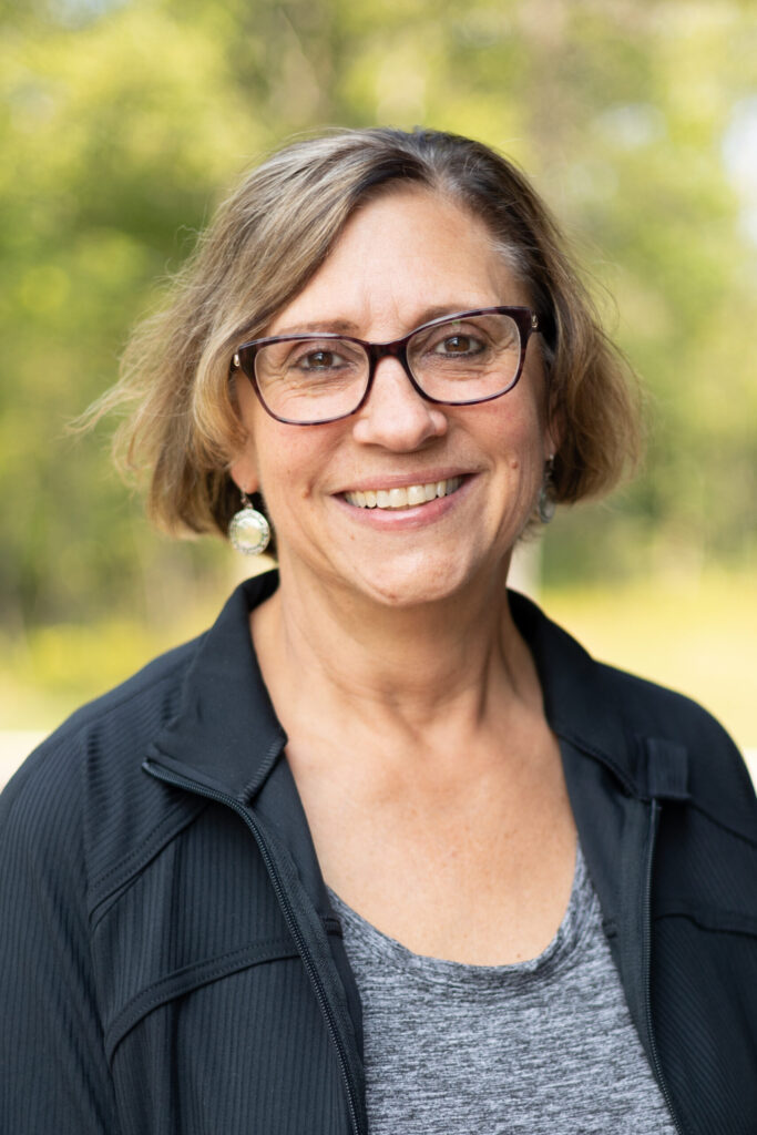 A smiling woman with short, light brown hair and glasses is wearing a dark jacket and gray top, standing outdoors with green trees blurred in the background.