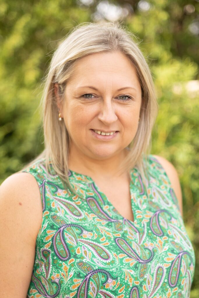 A woman with straight, blonde hair smiles softly at the camera. She is wearing a green sleeveless top with a colorful paisley pattern. The background is blurred greenery.