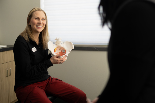 A healthcare professional smiles while holding and explaining a model of the human pelvis to a seated patient in a medical office.