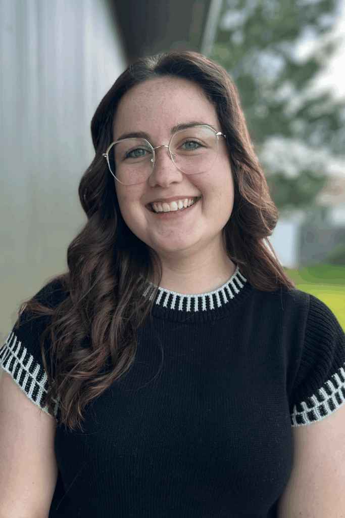 A young woman with long brown hair and glasses smiles at the camera. She is wearing a black shirt with white striped trim and is standing outside near a building, with greenery visible in the background.