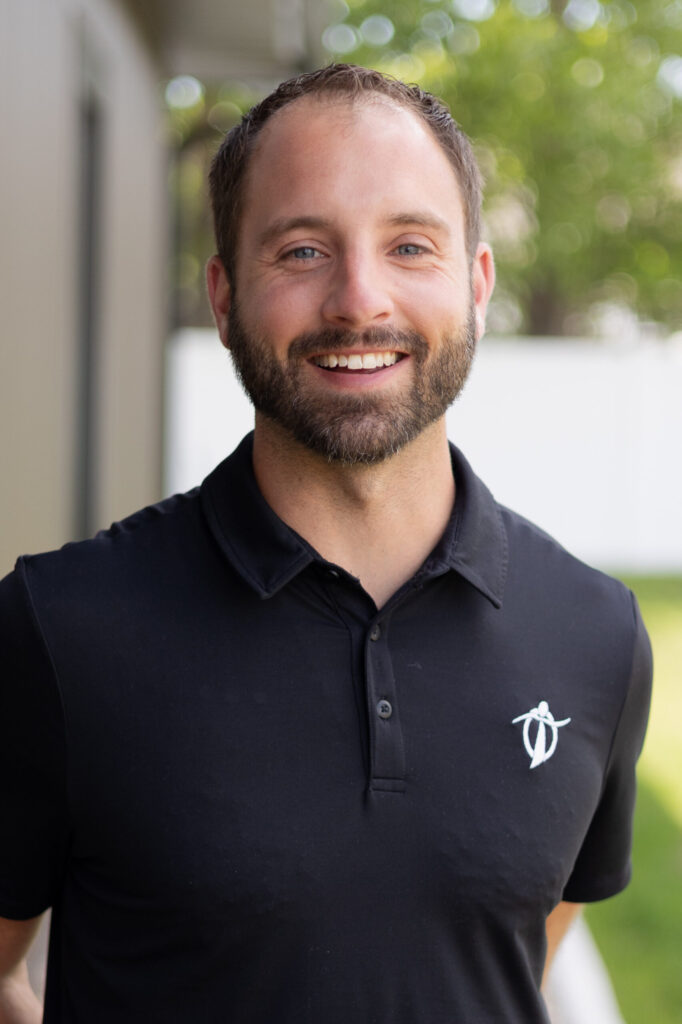 A smiling man with short brown hair and a beard wears a black polo shirt with a white logo, standing outdoors with greenery and a white fence in the background.