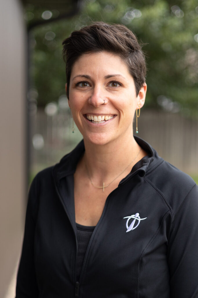 A smiling woman with short brown hair, wearing a black zip-up jacket with a dance logo, a gold necklace, and drop earrings, stands outdoors with greenery blurred in the background.