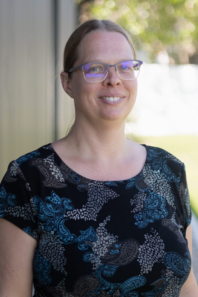 A woman with light skin, wearing glasses and a black top with a blue and white paisley pattern, stands outside, smiling. The background is blurred with greenery and a building visible.