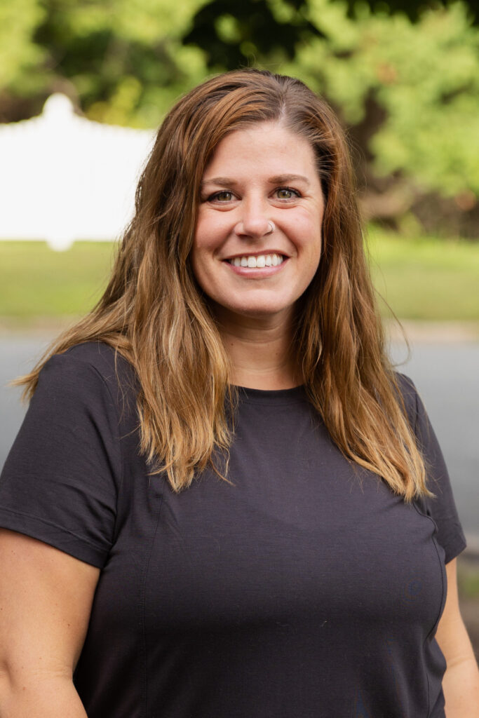 A woman with long, light brown hair wearing a black t-shirt smiles outdoors with trees and a white fence blurred in the background.