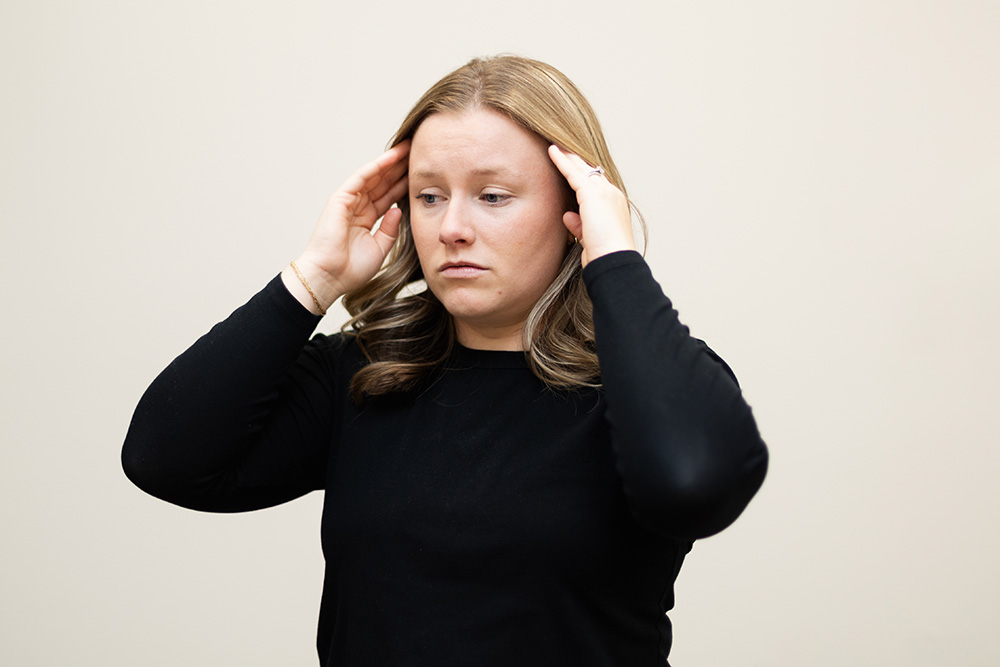 A woman with light brown hair wearing a black long-sleeve shirt touches her temples with both hands and looks down with a concerned or stressed expression. The background is plain and light-colored.