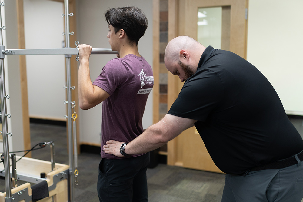 A physical therapist in a black shirt assists a young man in a purple shirt who is holding onto a metal bar, guiding his lower back during a physical therapy exercise in a clinic.