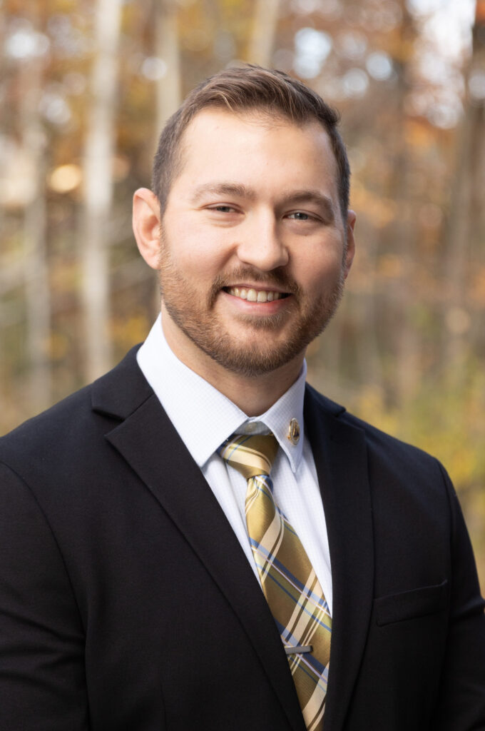 A man with short light brown hair and a beard is wearing a black suit jacket, white shirt, and a yellow plaid tie. He is smiling and standing outdoors with blurred trees in the background.