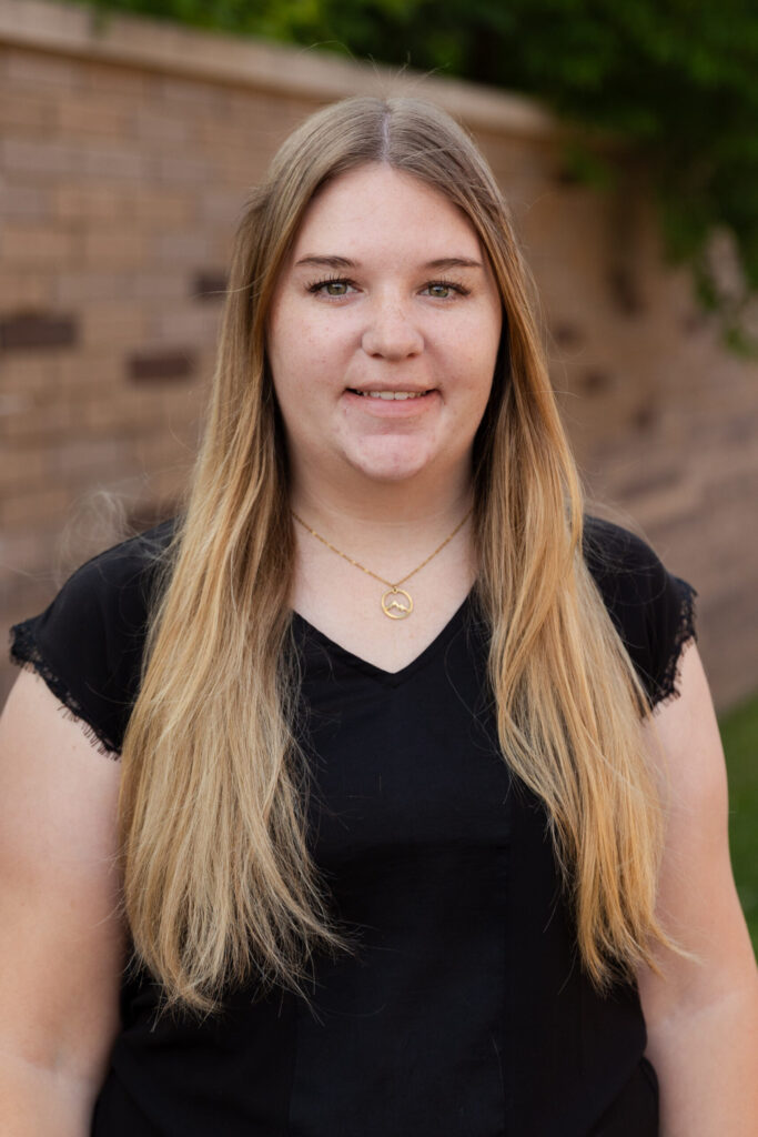 A young woman with long blonde hair wearing a black shirt and a gold necklace stands outdoors in front of a brick wall, smiling at the camera.