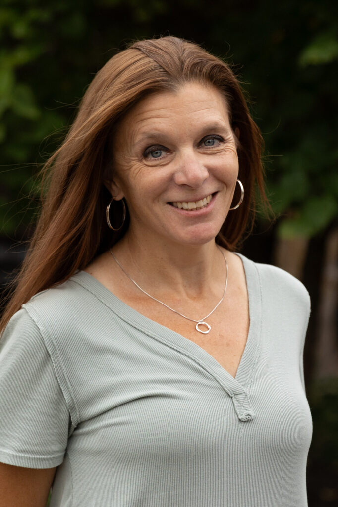 A woman with long brown hair, wearing a light green short-sleeve shirt, hoop earrings, and a silver necklace, smiles while standing outdoors with greenery in the background.