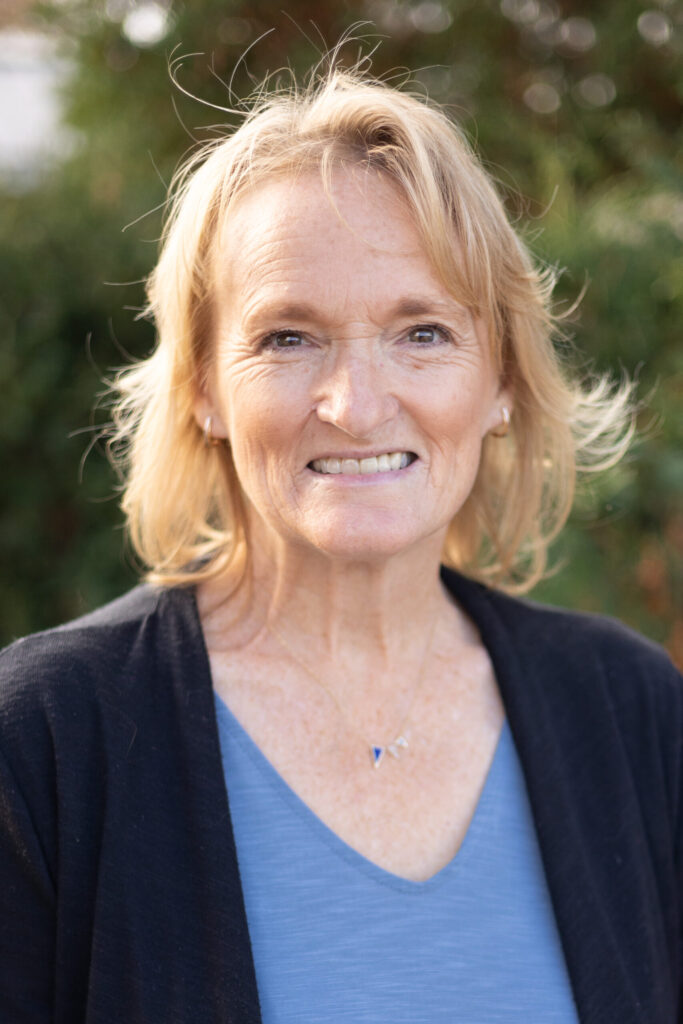 A smiling older woman with blonde hair wearing a blue top and black cardigan stands outdoors in front of green foliage.