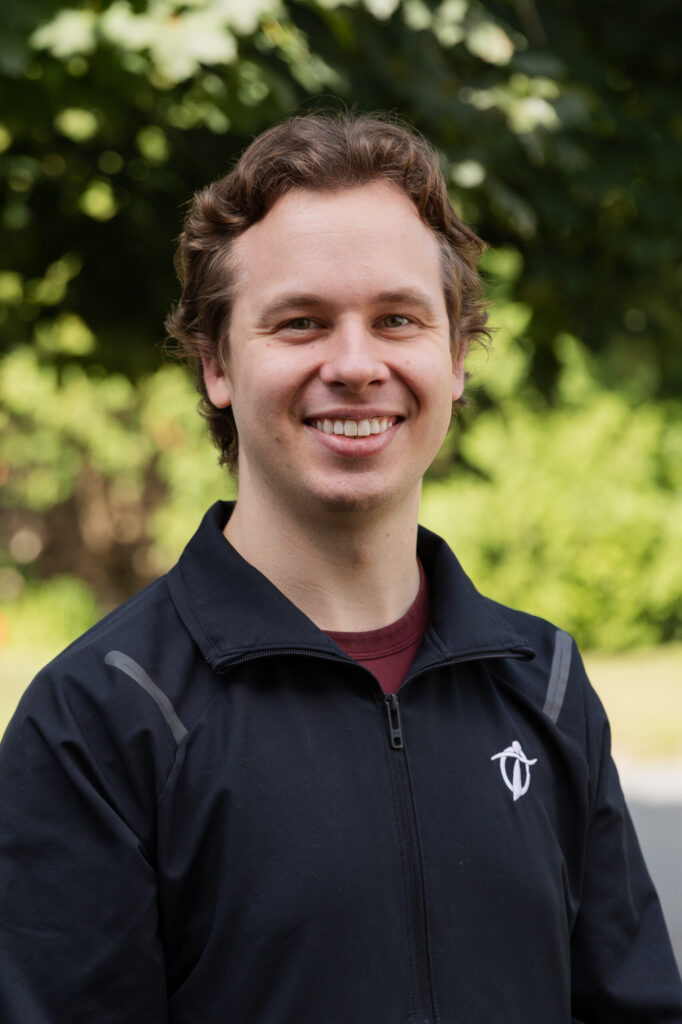 A young man with wavy brown hair smiles at the camera while standing outdoors. He is wearing a black jacket with a white logo and a maroon shirt, with green trees blurred in the background.