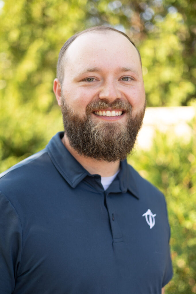 A bearded man with short hair, wearing a navy blue collared shirt with a white logo, smiles while standing outdoors, with green foliage in the background.