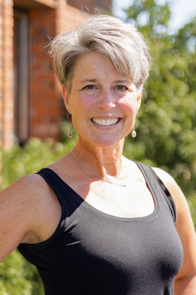 A smiling woman with short gray hair wearing a black sleeveless top and earrings stands outdoors near a brick wall, with green foliage and sunlight in the background.
