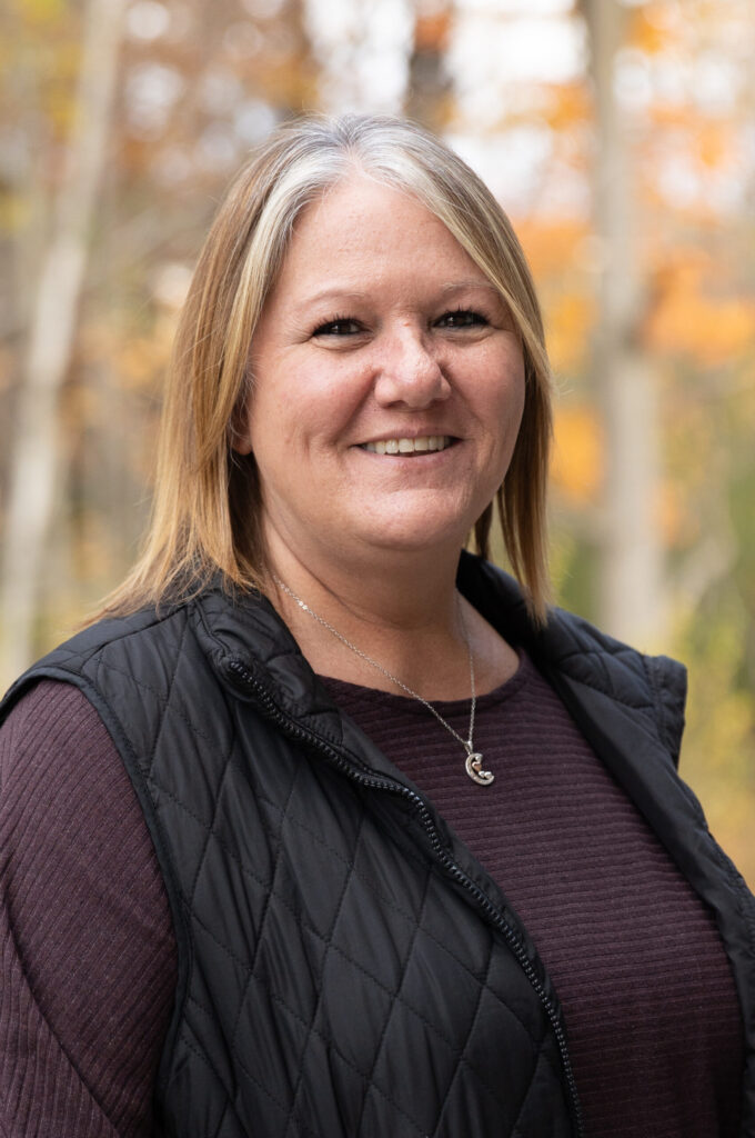 A woman with straight blonde hair, wearing a quilted black vest and a maroon top, smiles outdoors with blurred autumn trees in the background.