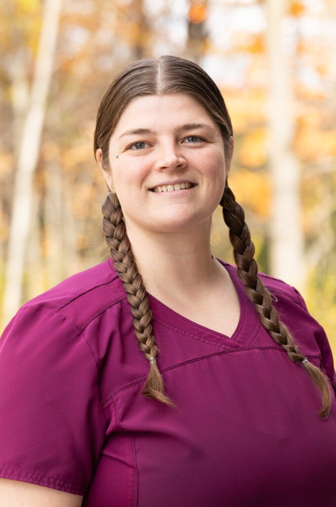 A woman with long brown hair in two braids, wearing a magenta scrub top, smiles at the camera. The background features soft-focus autumn trees with yellow and orange leaves.