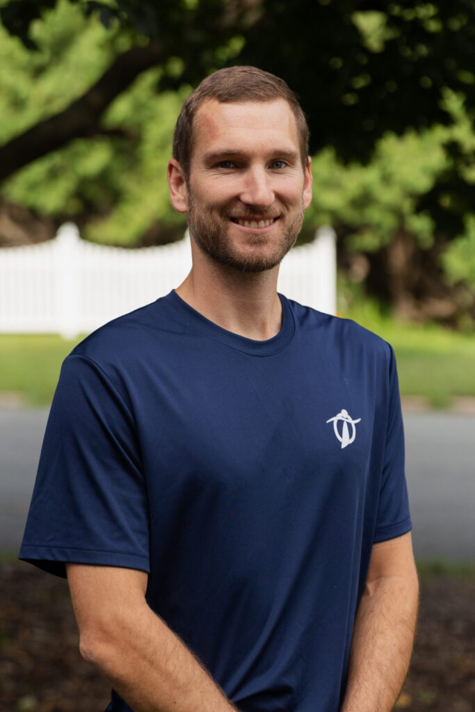 A man with short light brown hair and a beard, wearing a navy blue T-shirt with a white logo, stands outdoors smiling with trees and a white fence in the background.