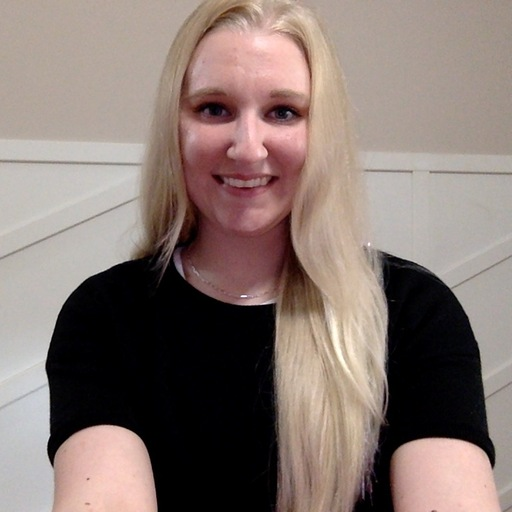 A woman with long blonde hair, wearing a black shirt, smiles at the camera in a well-lit room with light-colored walls and white paneling.
