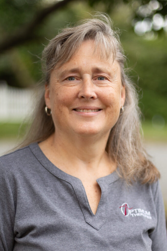 A middle-aged woman with long gray hair and blue eyes smiles outdoors, wearing a gray shirt with a logo that reads OPTIMA on the left side. The background is blurred with greenery and a white fence.