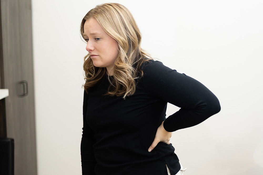 A woman with wavy blonde hair, wearing a black long-sleeve shirt, stands indoors with a concerned expression, holding her lower back with one hand as if experiencing discomfort.