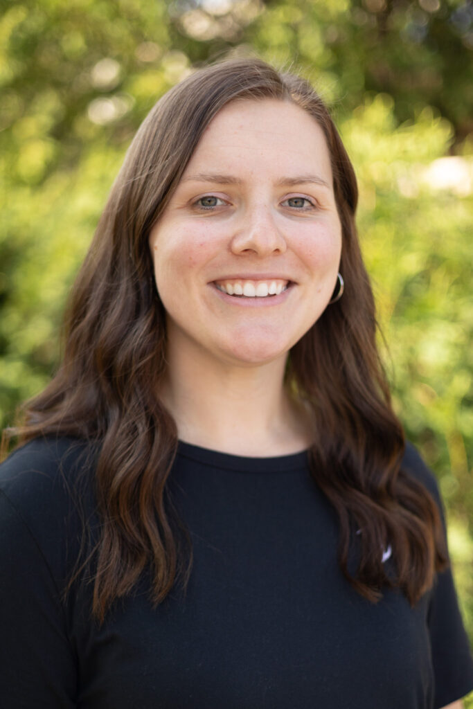A woman with long brown hair and a black shirt smiles at the camera while standing outside with greenery blurred in the background.
