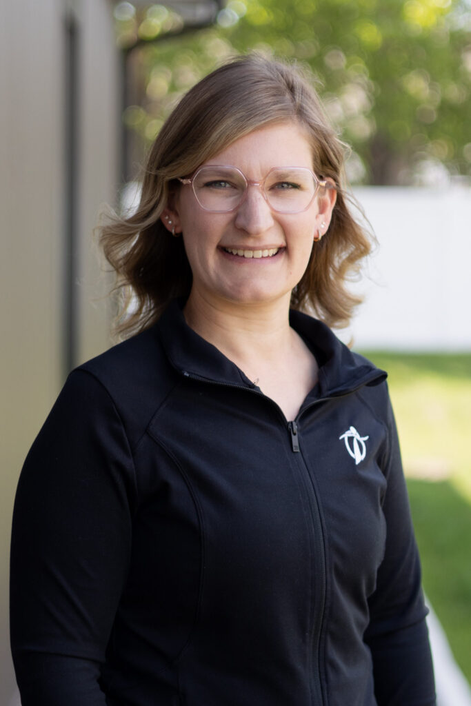 A woman with shoulder-length light brown hair and glasses smiles outdoors. She is wearing a black zip-up jacket with a white logo on the left side. The background has greenery and a white fence.