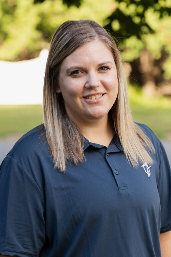 A woman with straight, shoulder-length blonde hair is smiling slightly. She is wearing a dark blue polo shirt and standing outdoors, with greenery and sunlight in the background.