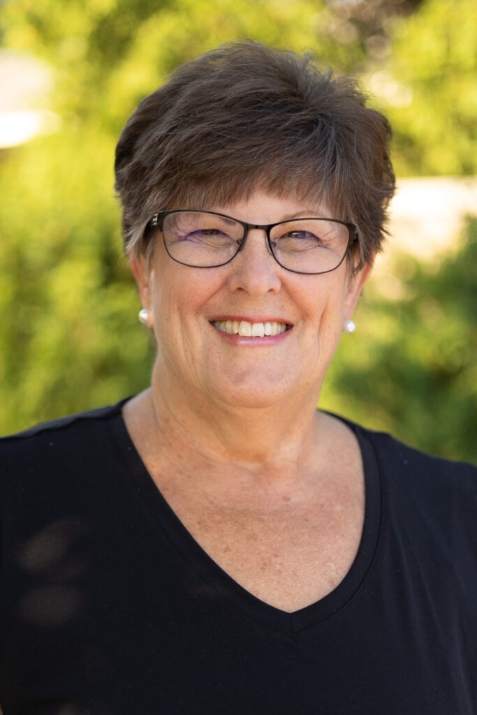A smiling older woman with short brown hair, glasses, and a black top stands outdoors in front of blurred green foliage.