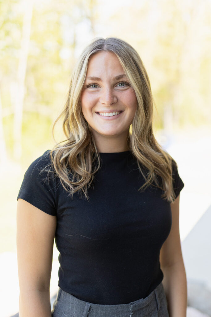 A young woman with long, wavy blonde hair, wearing a black short-sleeve top and gray pants, smiles at the camera while standing outdoors with a blurred background of trees.