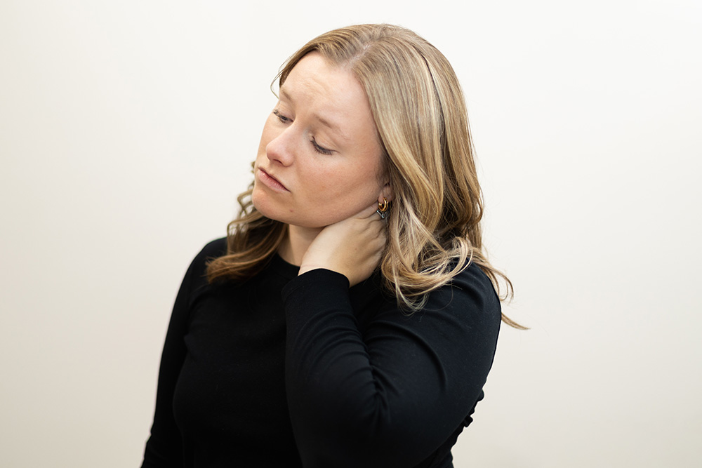 A woman with light brown hair wearing a black shirt holds her hand to the back of her neck and tilts her head, appearing to experience neck pain or discomfort. The background is plain and light-colored.