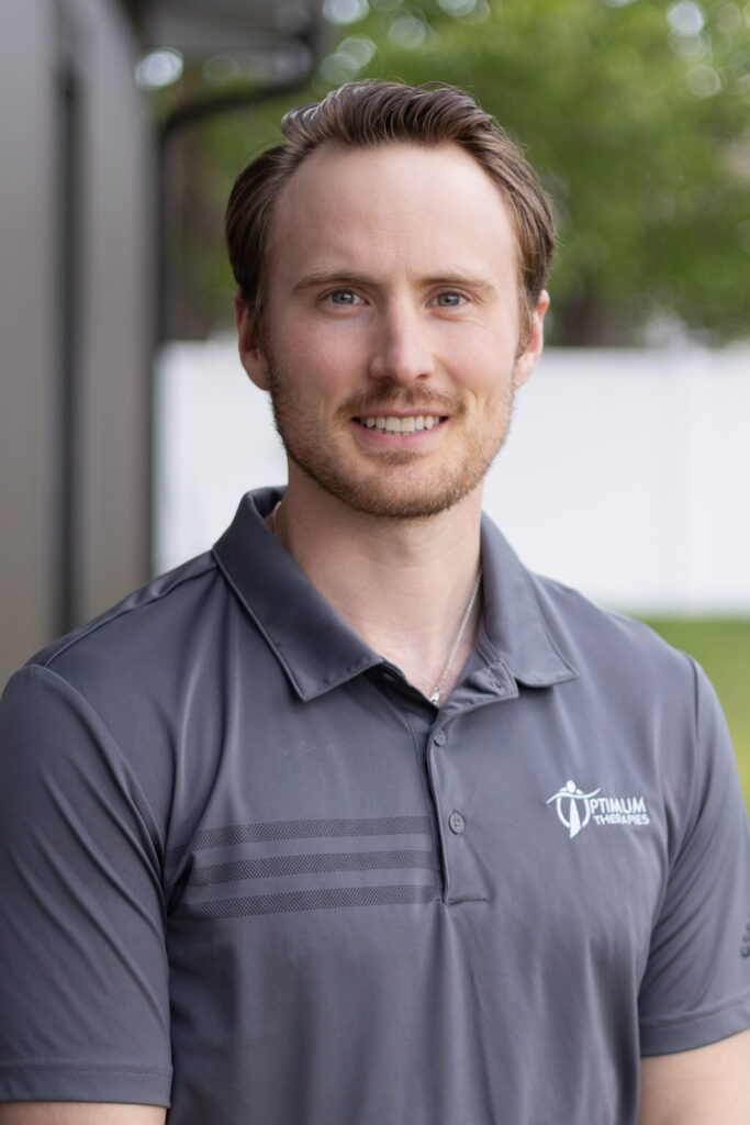 A man with light brown hair and a trimmed beard smiles at the camera, wearing a gray polo shirt with the Optimum Vitality logo. The background is outdoors with blurred greenery and a white fence.