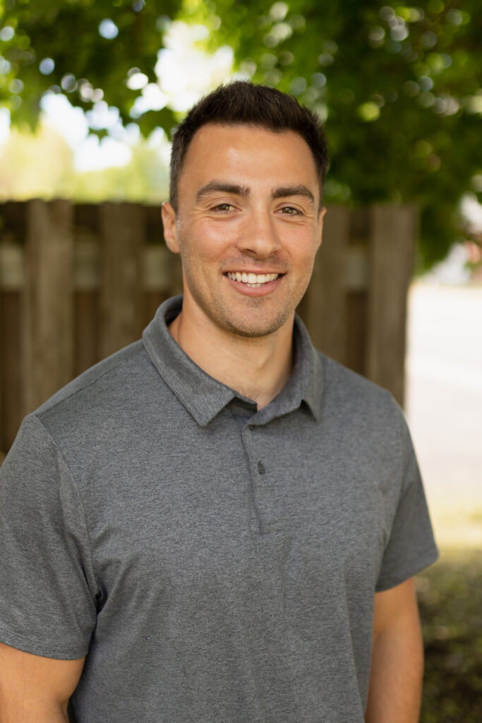 A man with short dark hair wearing a gray polo shirt smiles while standing outdoors in front of a wooden fence and leafy green trees.