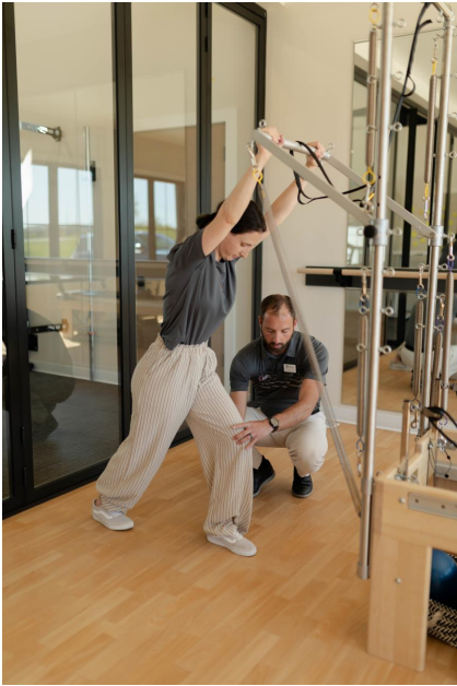 A woman performs a stretching exercise using resistance bands on a Pilates machine while a male trainer kneels beside her, guiding her posture in a bright, modern studio.