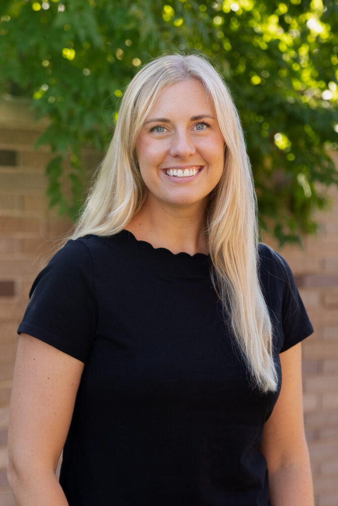 A smiling woman with long blonde hair wearing a black short-sleeved top stands outdoors in front of a leafy green tree and a brick wall.