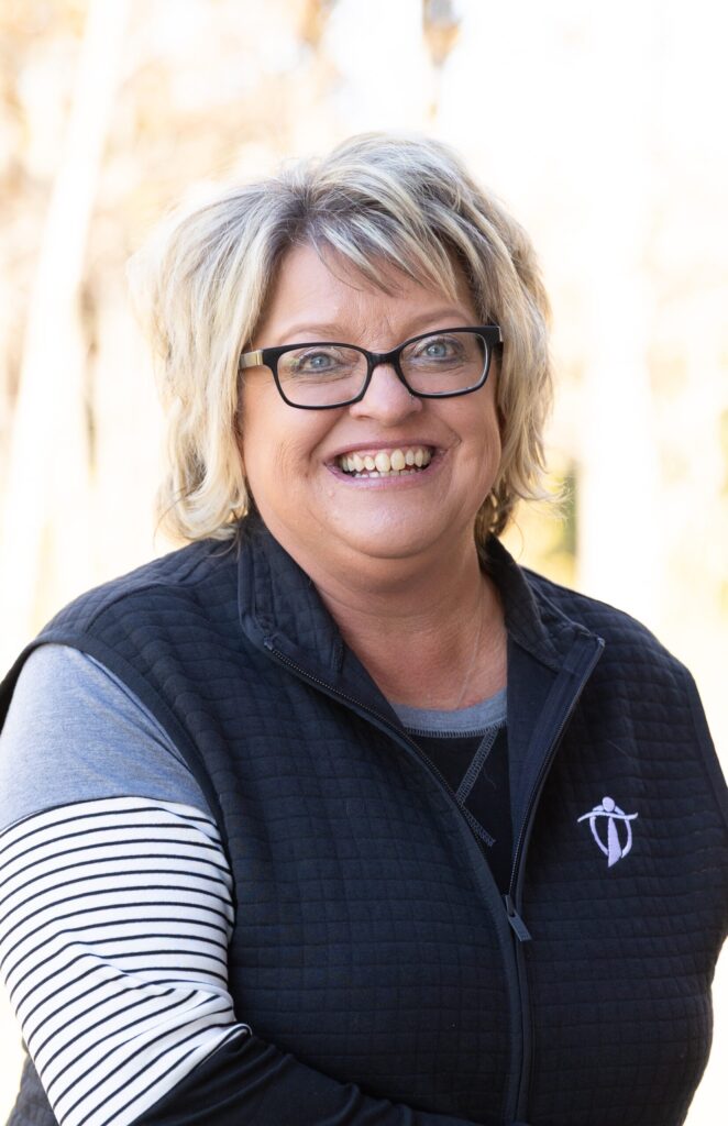 A smiling woman with short blonde hair and glasses is wearing a black quilted vest over a striped long-sleeve shirt, standing outdoors with a blurred, sunlit background.