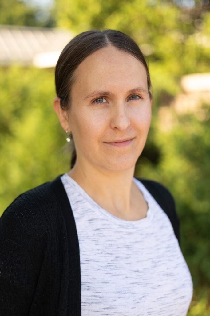 A woman with straight brown hair pulled back, wearing a white top and black cardigan, stands outdoors with a soft smile. Green foliage and blurred sunlight are visible in the background.