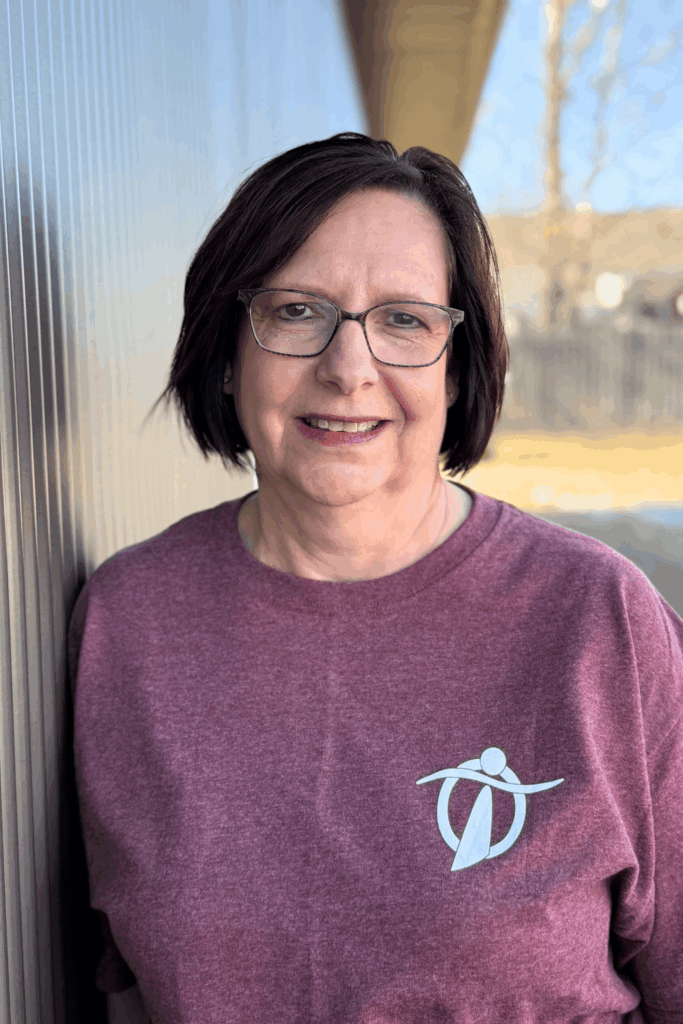 A woman with short dark hair and glasses smiles while standing outside against a wall. She is wearing a maroon T-shirt with a stylized white logo on the left side. The background is slightly blurred.