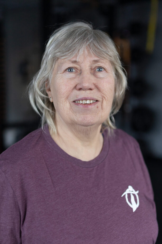 An older woman with gray hair and a purple shirt smiles softly at the camera. The background is softly blurred with dark tones, drawing focus to her relaxed expression.