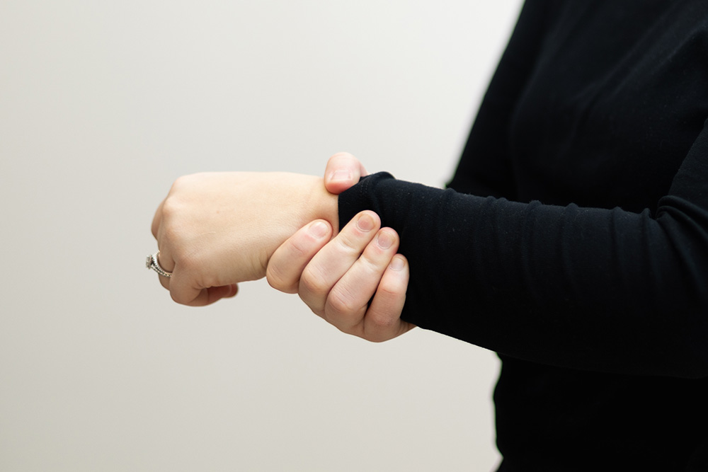 A person wearing a black long-sleeve shirt adjusts their left sleeve with their right hand against a plain light background.
