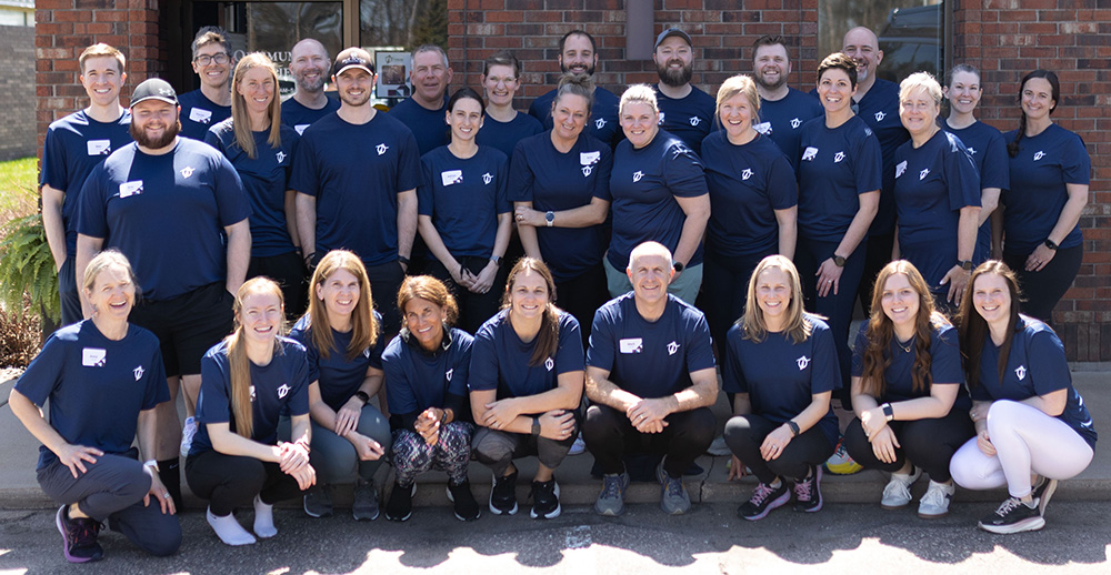 A group of about 30 people wearing matching navy blue shirts pose together outside in front of a brick building, smiling at the camera. Some are standing, while others crouch or kneel in front.
