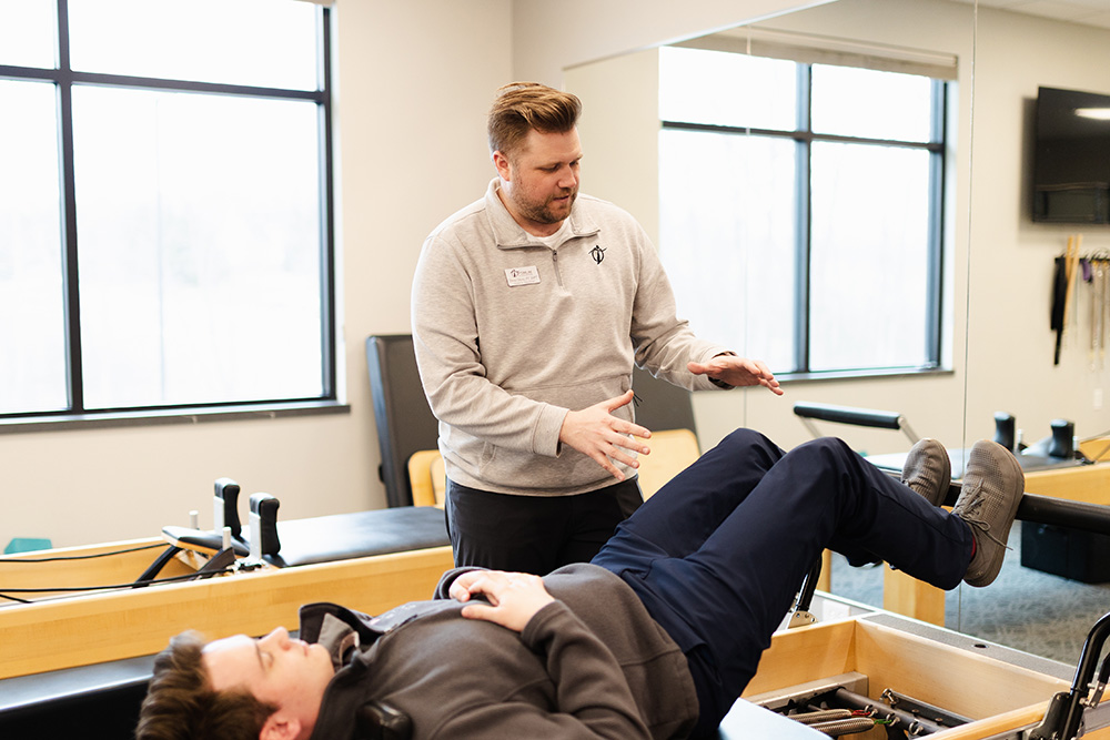 A physical therapist guides a patient through a leg exercise on a rehabilitation table in a bright, modern clinic with large windows.