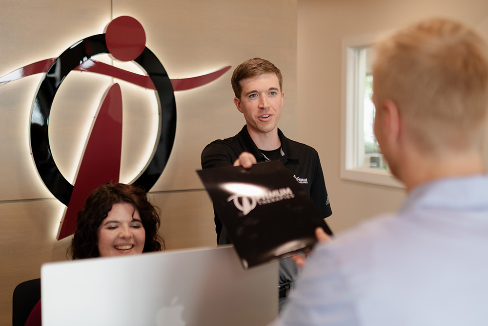 A man hands a folder to another man at a reception desk, while a woman sits smiling behind an Apple computer. A large, stylized logo is displayed on the wall in the background.